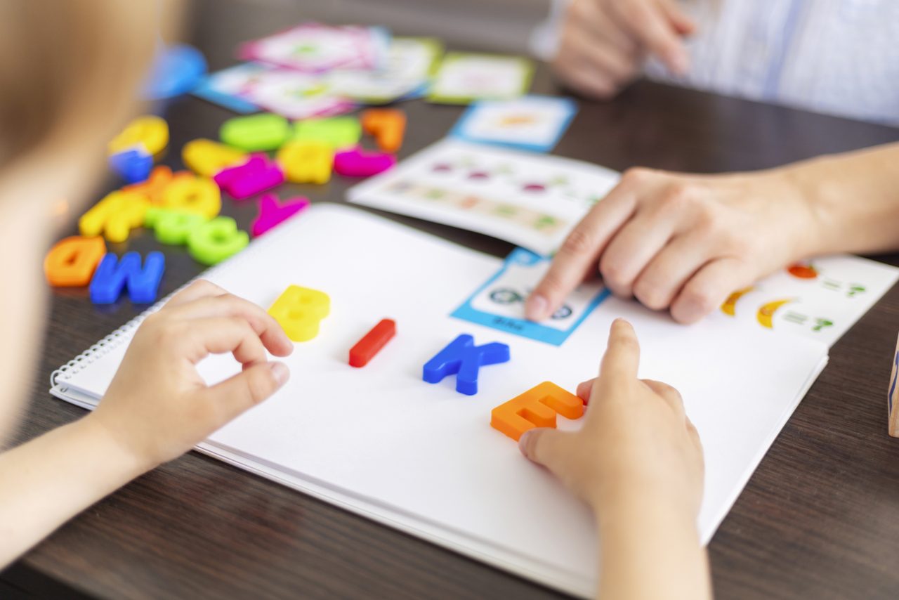 Child with dyslexia practicing reading using multisensory tools—tracing letters in colored sand, blending sounds with magnetic tiles, and reading a decodable book with a tutor